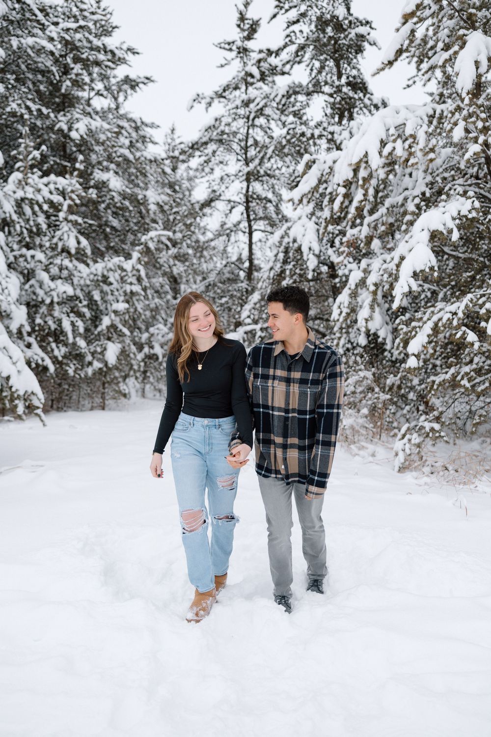 A couple walks hand in hand through a snow-covered forest landscape.