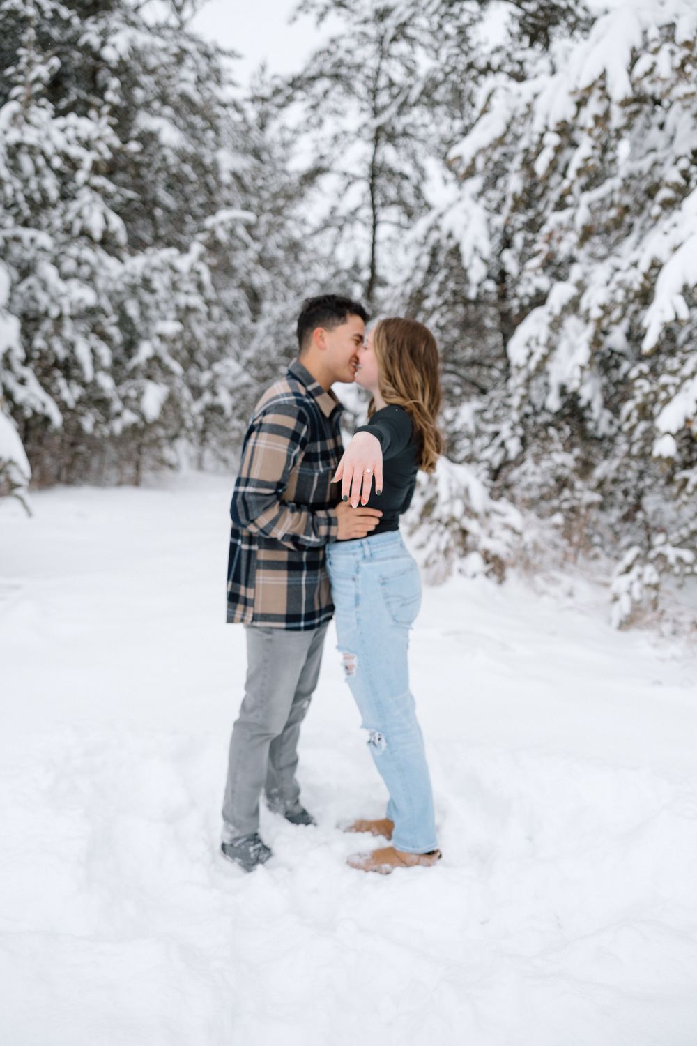 A romantic winter photoshoot with a couple in casual attire against snow-covered trees.
