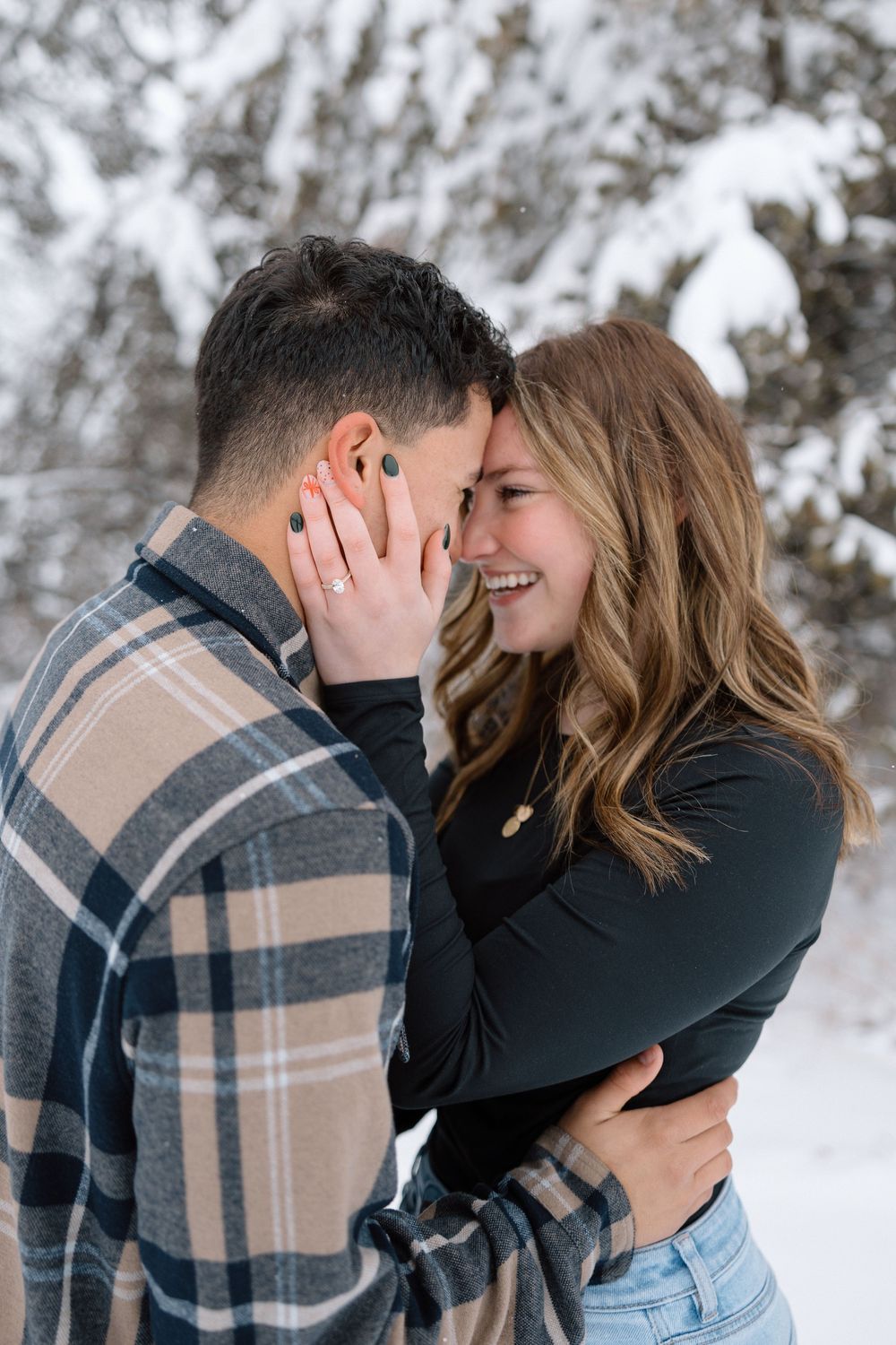Romantic winter photoshoot with couple in plaid and black embracing in snowy forest setting.