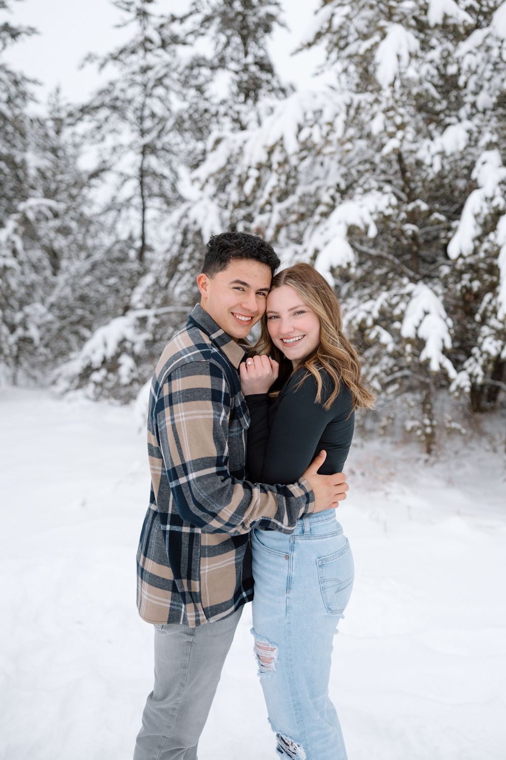 Playful couple laughing together against snowy winter backdrop wearing casual outfits.