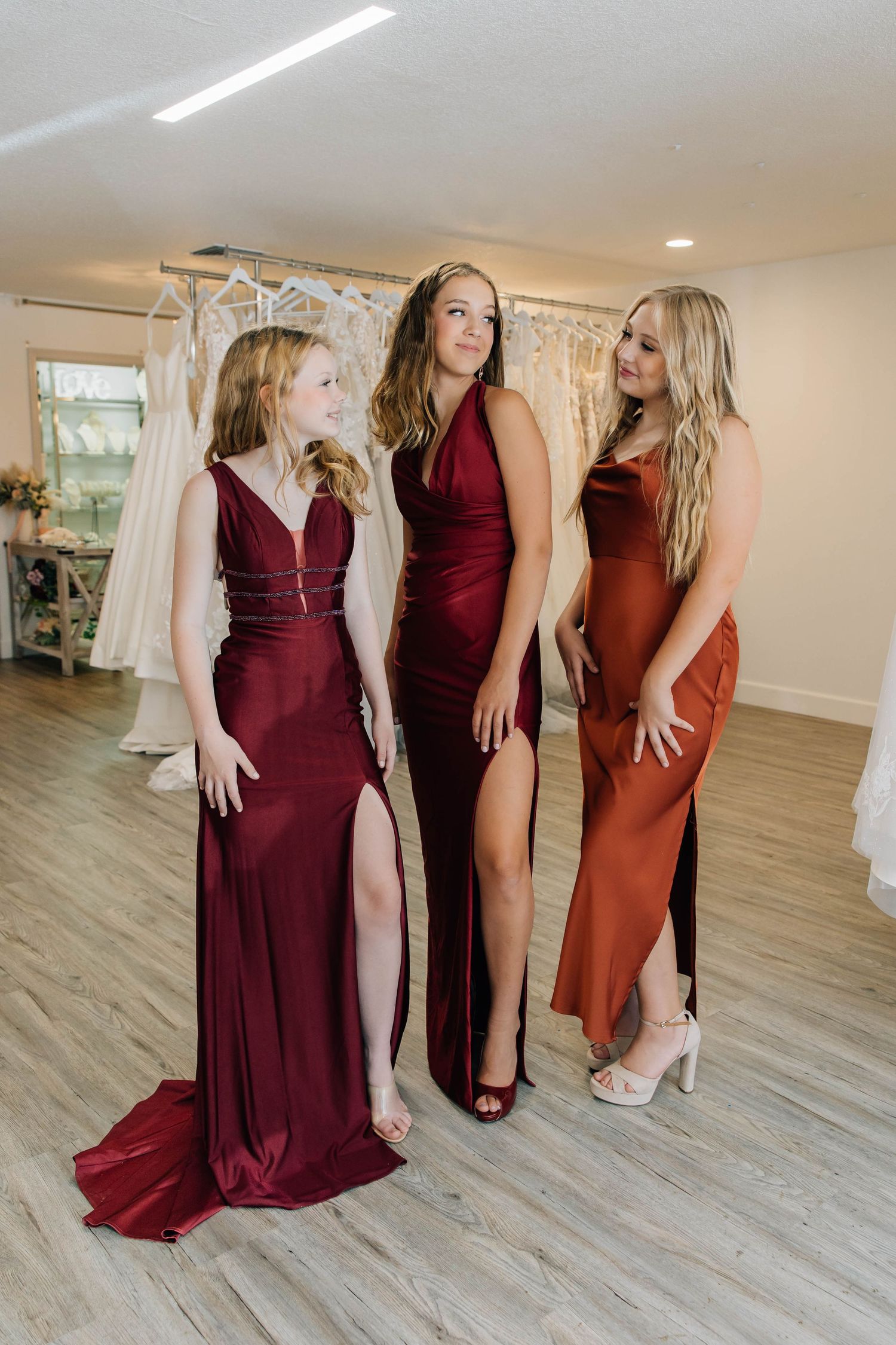 Three young women in elegant formal gowns pose together in a bridal boutique.