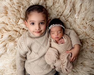 Two siblings in cream sweaters pose together on fluffy white rug for professional portrait.