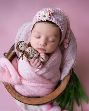 Sleeping newborn wrapped in pink knit blanket with matching bonnet and vintage toy in wooden bowl.