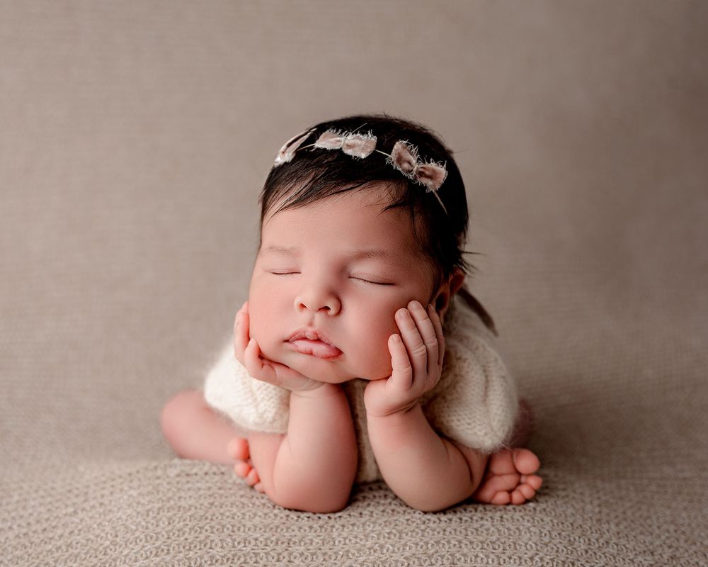 Sleeping baby posed with hands under cheeks wearing delicate headband on neutral background.