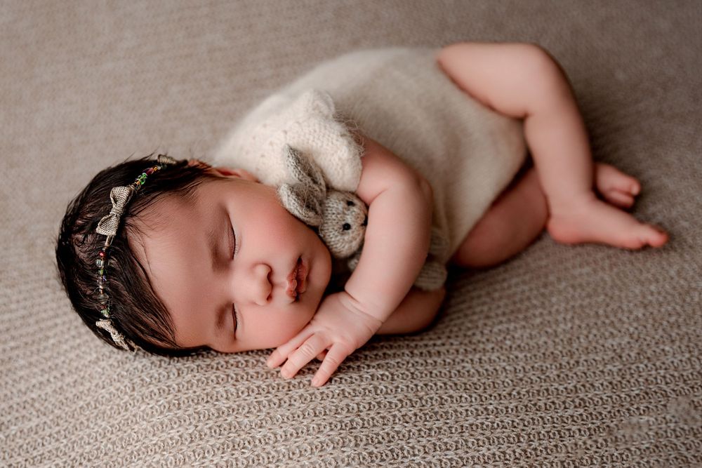 Sleeping infant in lace headband and white wrap on neutral textured background.