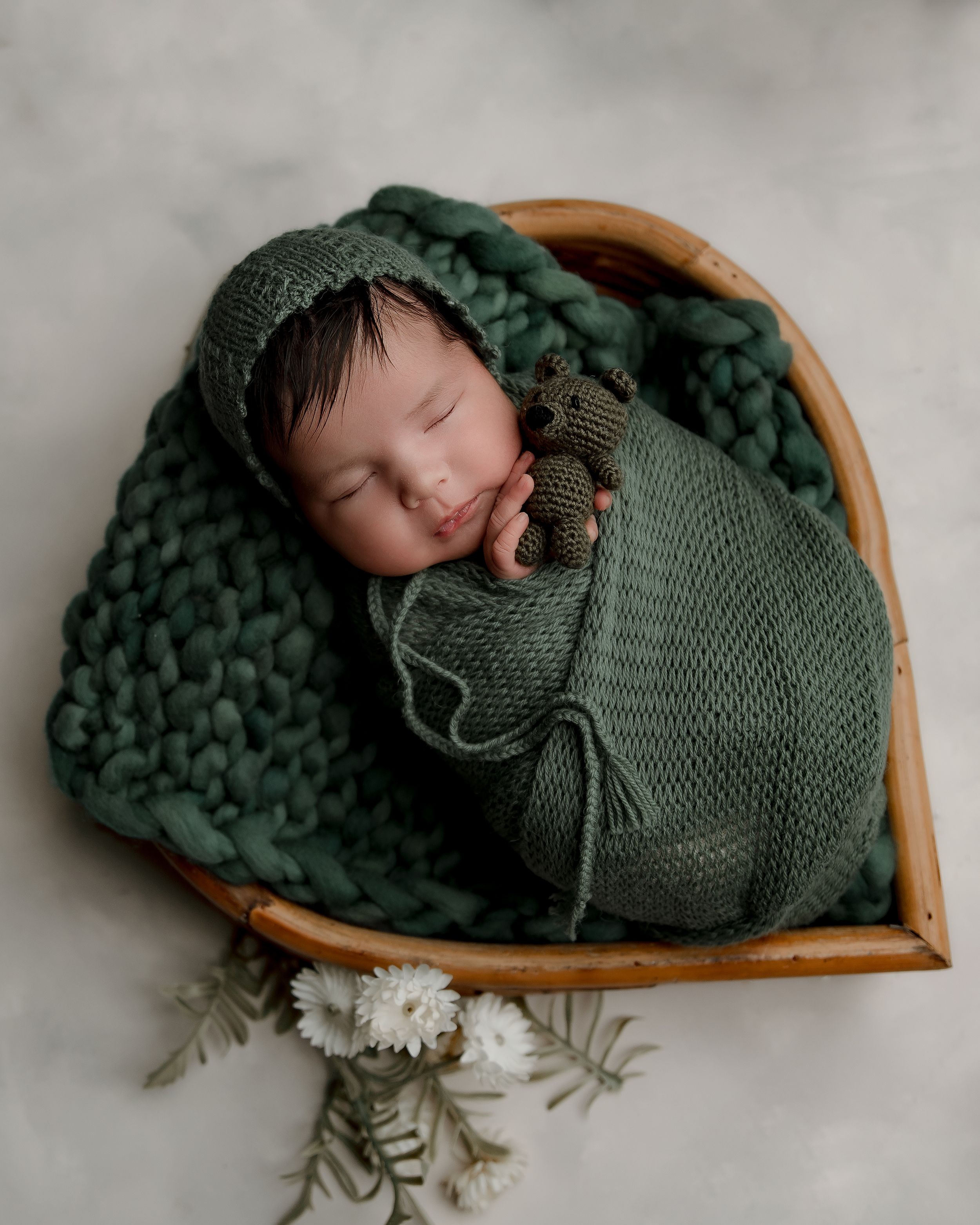 Peaceful newborn sleeping in wooden heart basket wrapped in deep green knitted blanket.