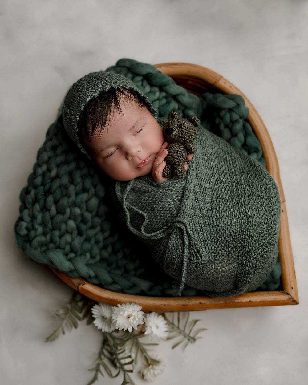 Peaceful newborn sleeping in wooden heart basket wrapped in deep green knitted blanket.
