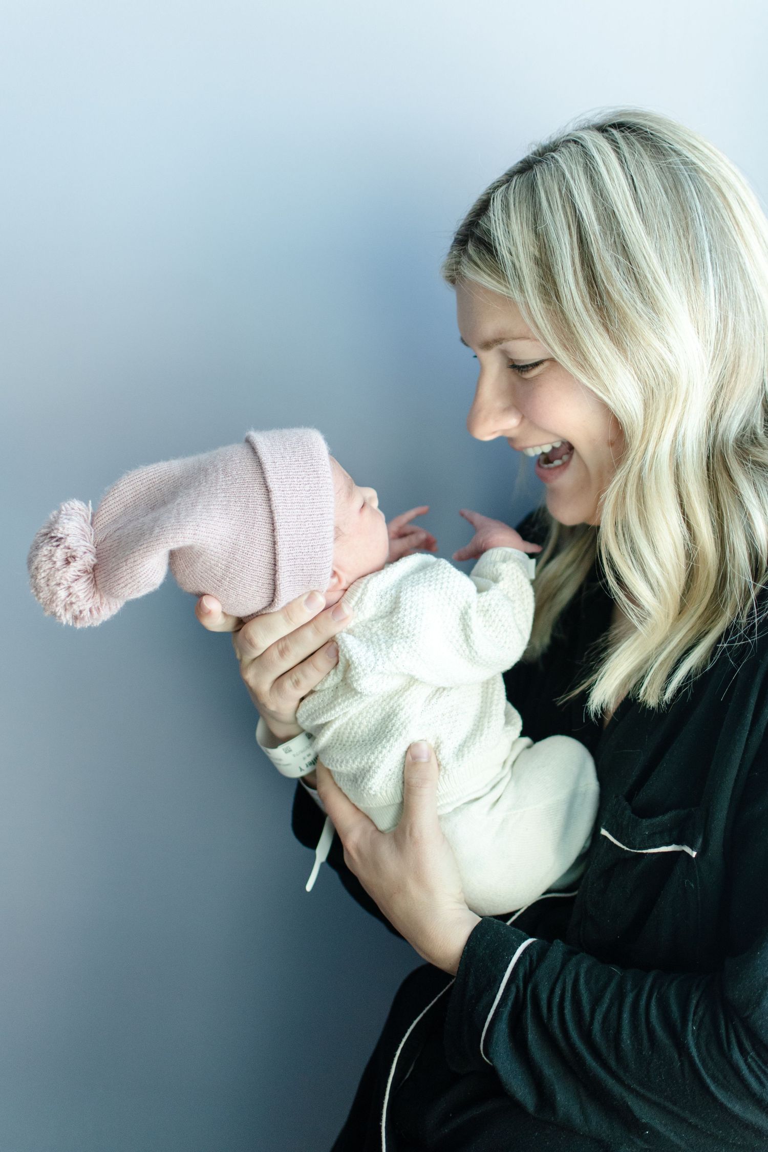 Woman in black holding laughing newborn wearing pink knit hat against grey background.