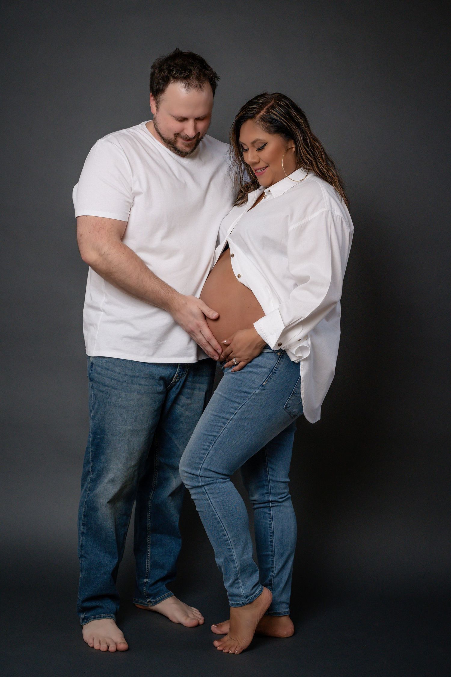 Intimate maternity photo shoot showing couple's feet and legs in casual white shirts and jeans against dark backdrop.