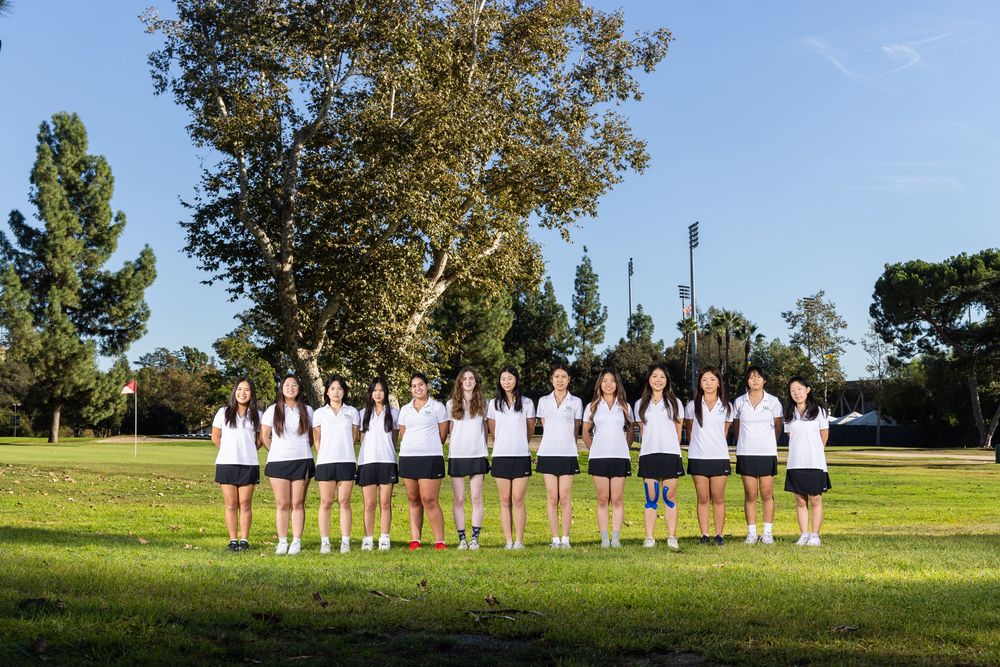A line of people in white shirts and dark shorts standing with their backs to camera on a sunny grass field.