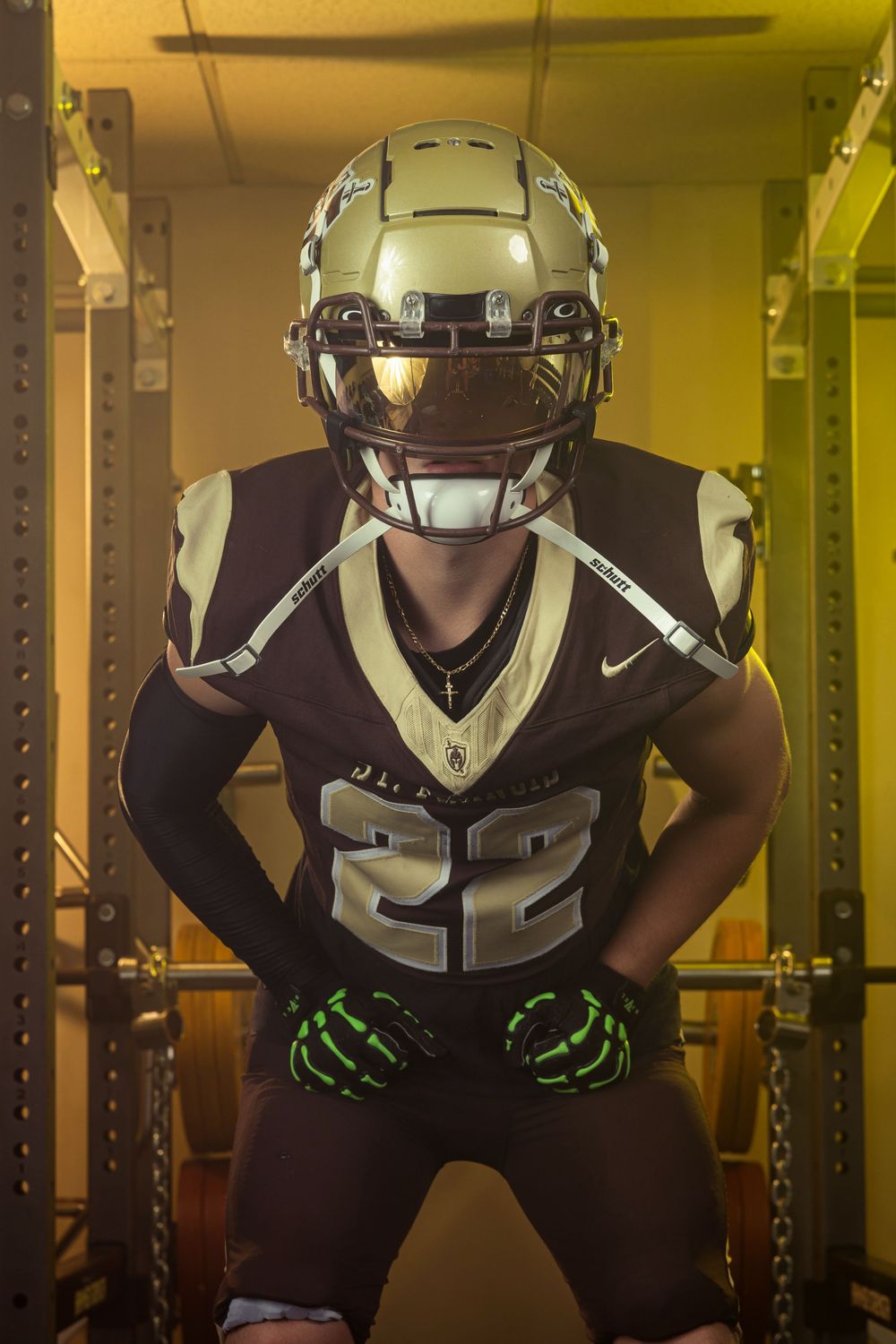 Football player #22 in dark uniform and gold helmet poses in weight room under dramatic lighting.