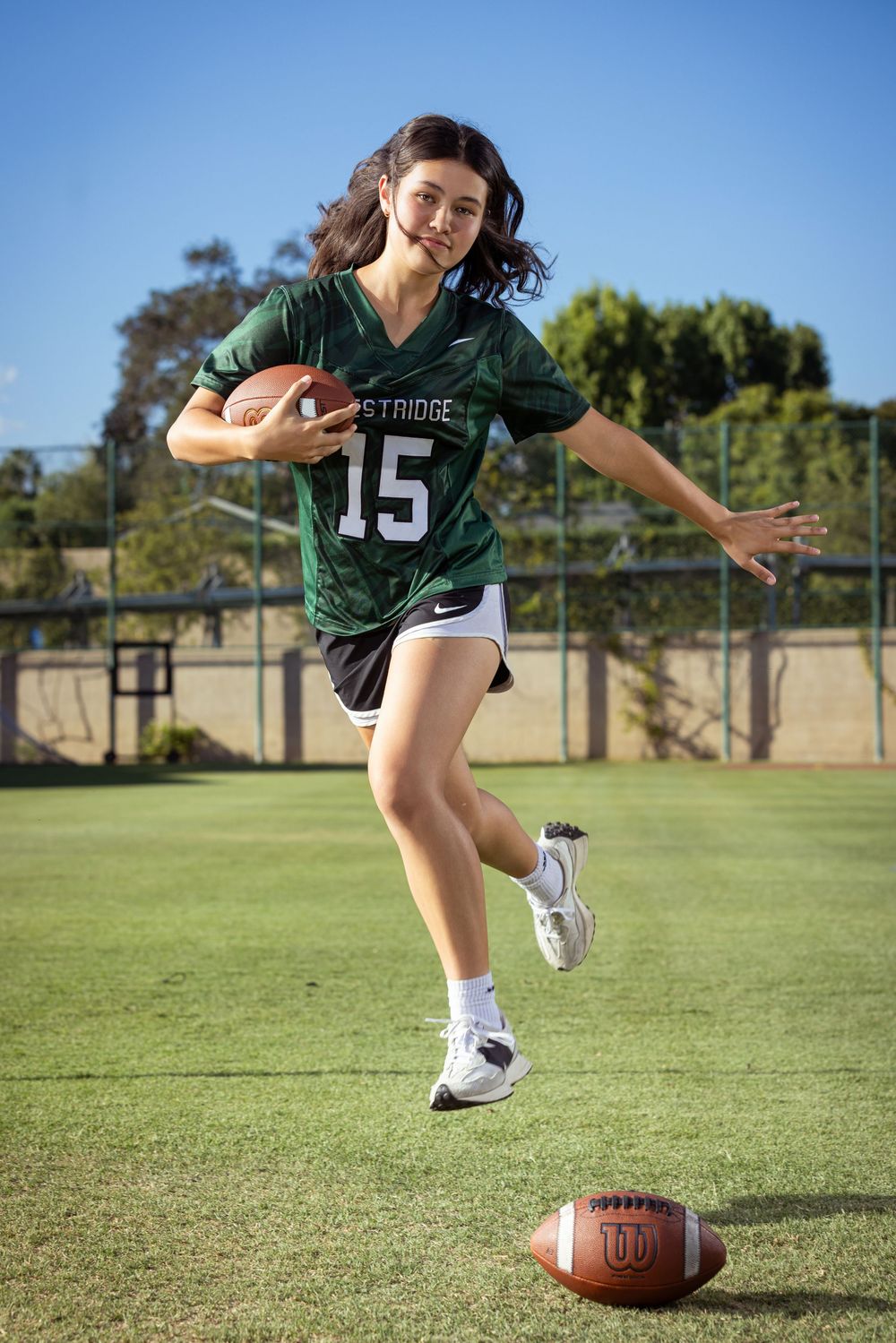 Player in green jersey number 15 runs with football on grass field during practice on sunny day.