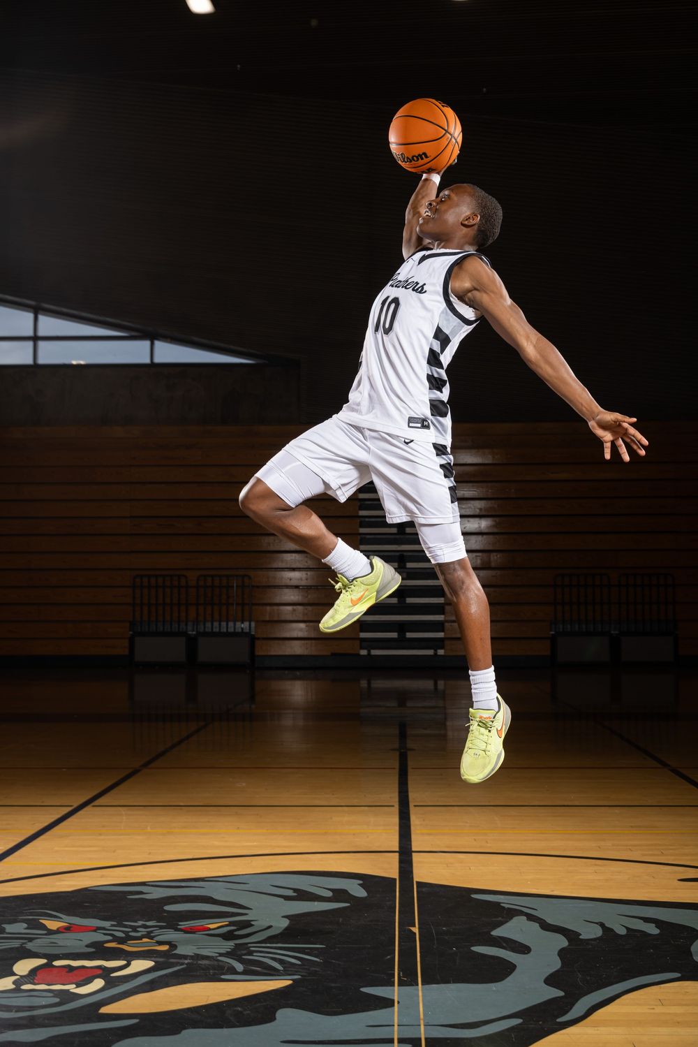 Basketball player leaps high in white uniform on dark court with mascot logo, demonstrating impressive vertical jump ability.