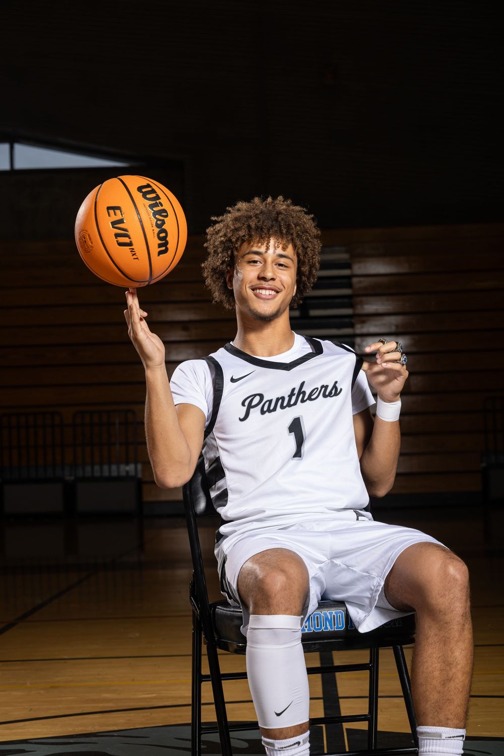 Basketball player in white Panthers jersey sits on chair holding orange ball in dark gymnasium.