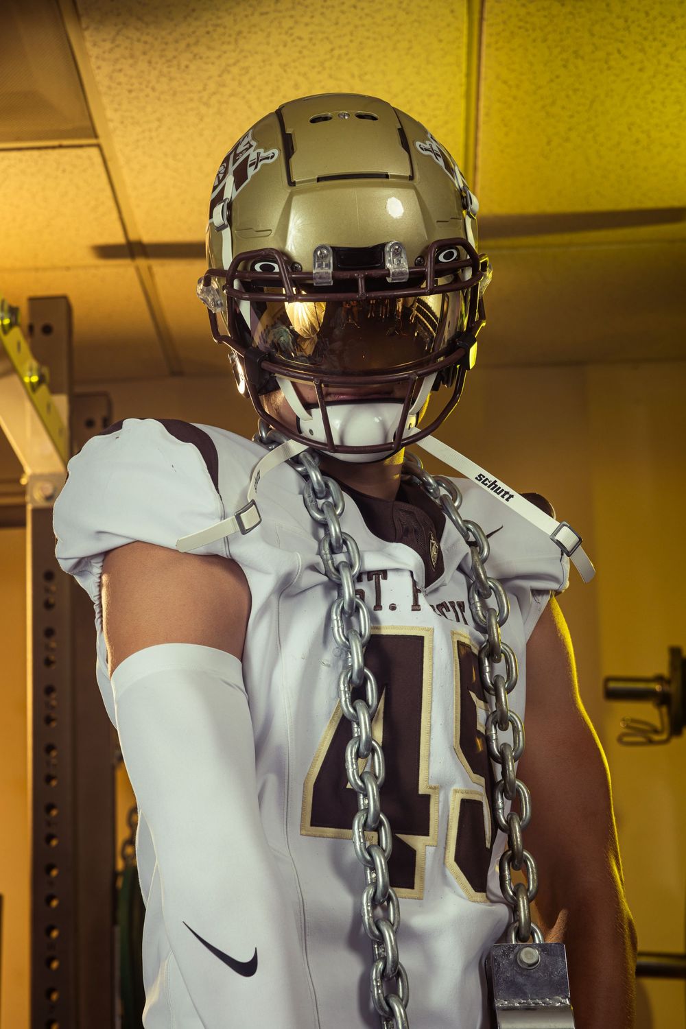 Football player in gold helmet and white Nike jersey wearing chains poses in locker room.