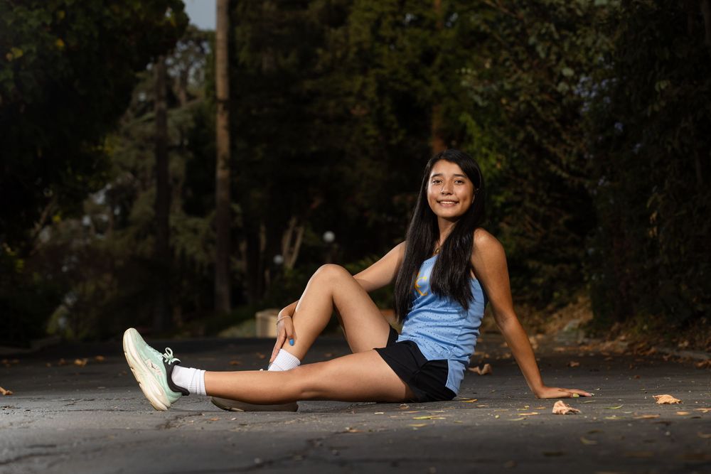 Young woman in blue tank top and black shorts sitting on a road, smiling, with trees in the background.