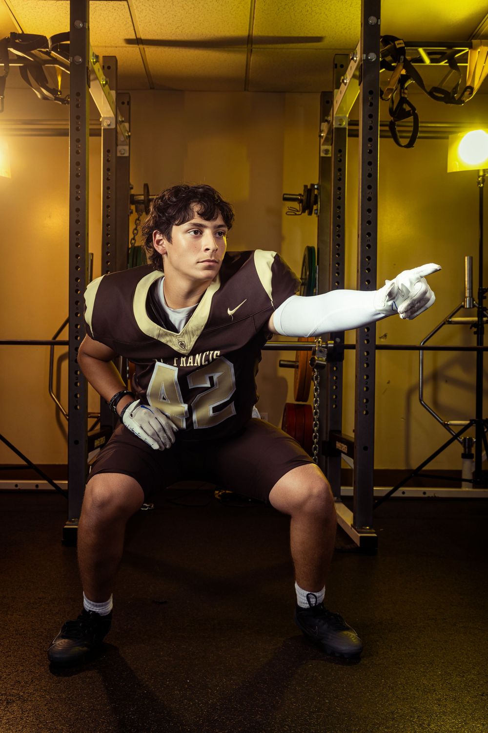 Young football player in brown jersey and gloves poses in a gym weight room under warm lighting.