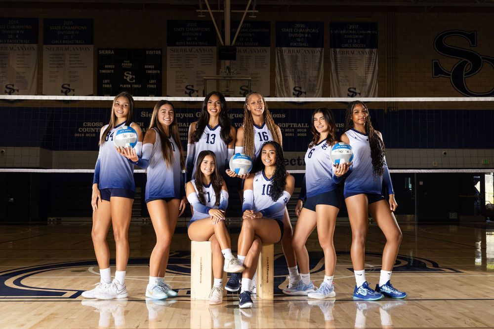 A volleyball team poses together in navy blue and white uniforms on a gymnasium court with a volleyball net behind them.