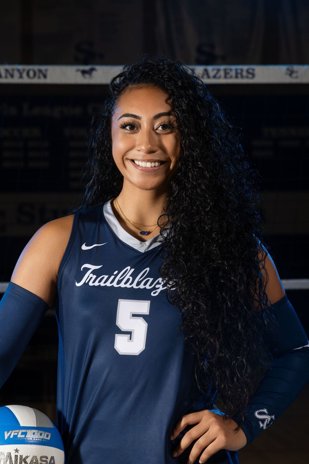 Smiling volleyball player wearing a Trailblazers number 5 jersey poses with a Mikasa ball in a gym.
