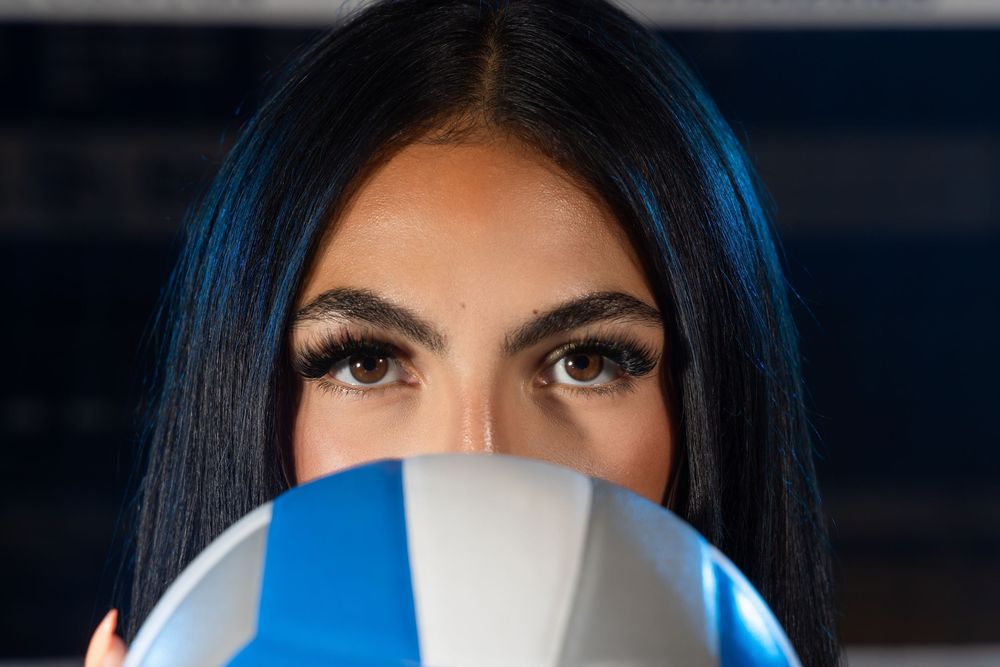Young woman with dark hair holding a blue and white volleyball in front of her face, showing expressive eyes.