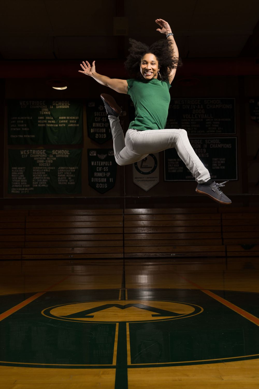 A person in a green shirt and white pants performs a dynamic dance move in a dimly lit gymnasium with wooden floors.