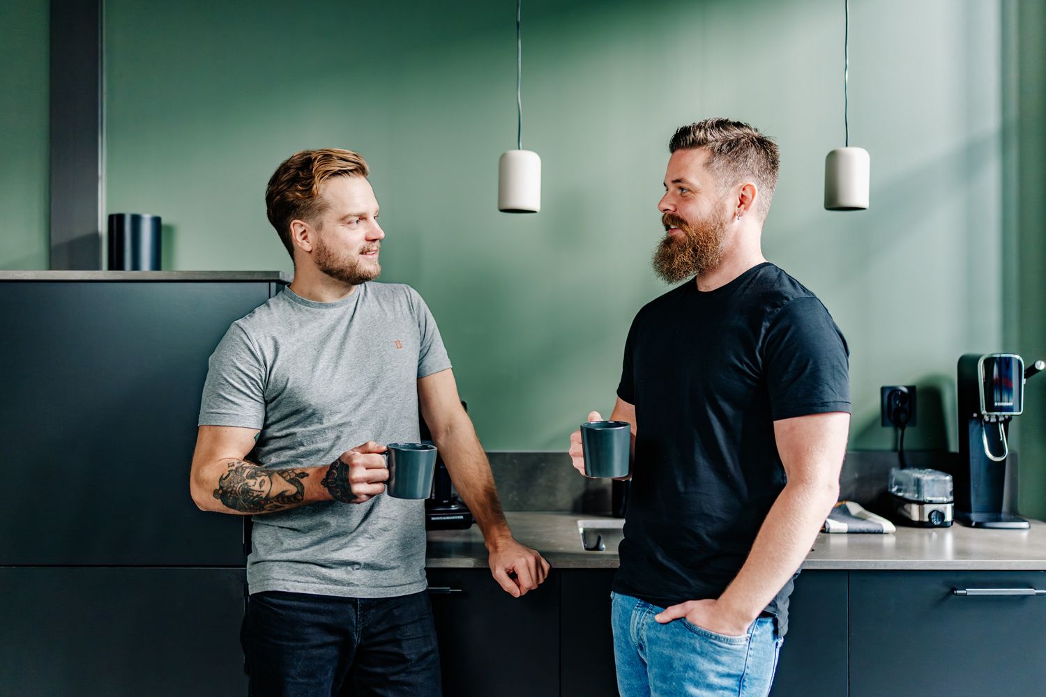 Två män står och pratar i ett kök med varsin kaffekopp i handen.
