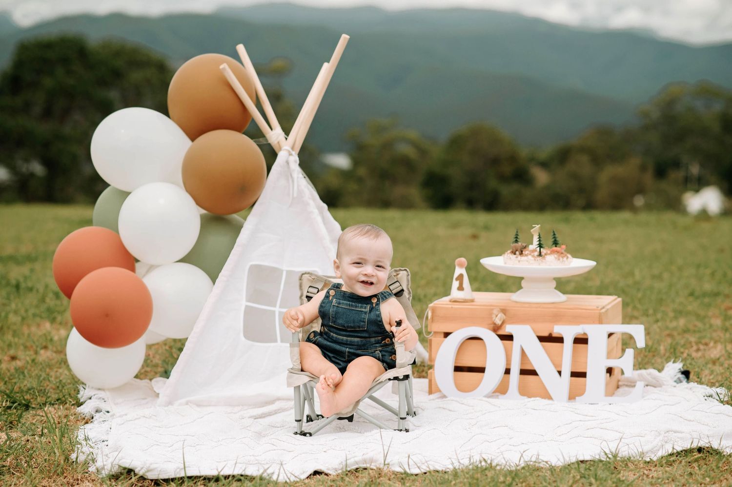 Baby in blue overalls sitting by teepee tent with ONE sign and birthday decorations.