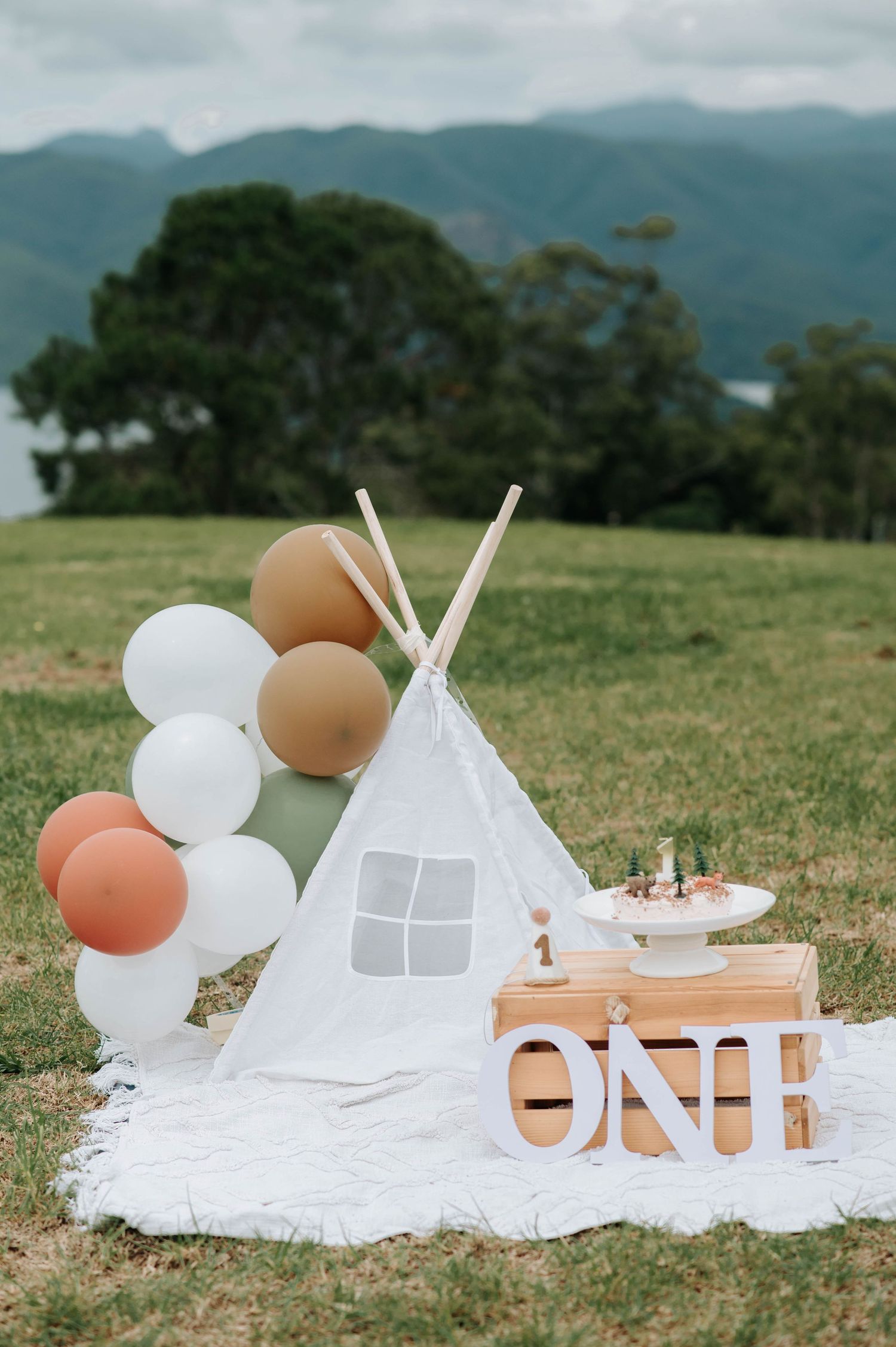 Outdoor first birthday setup with white teepee, balloon arch and ONE sign against mountain landscape.