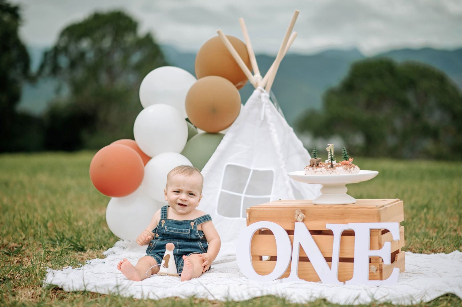 First birthday setup with teepee tent, balloons, and ONE sign on white blanket in outdoor setting.