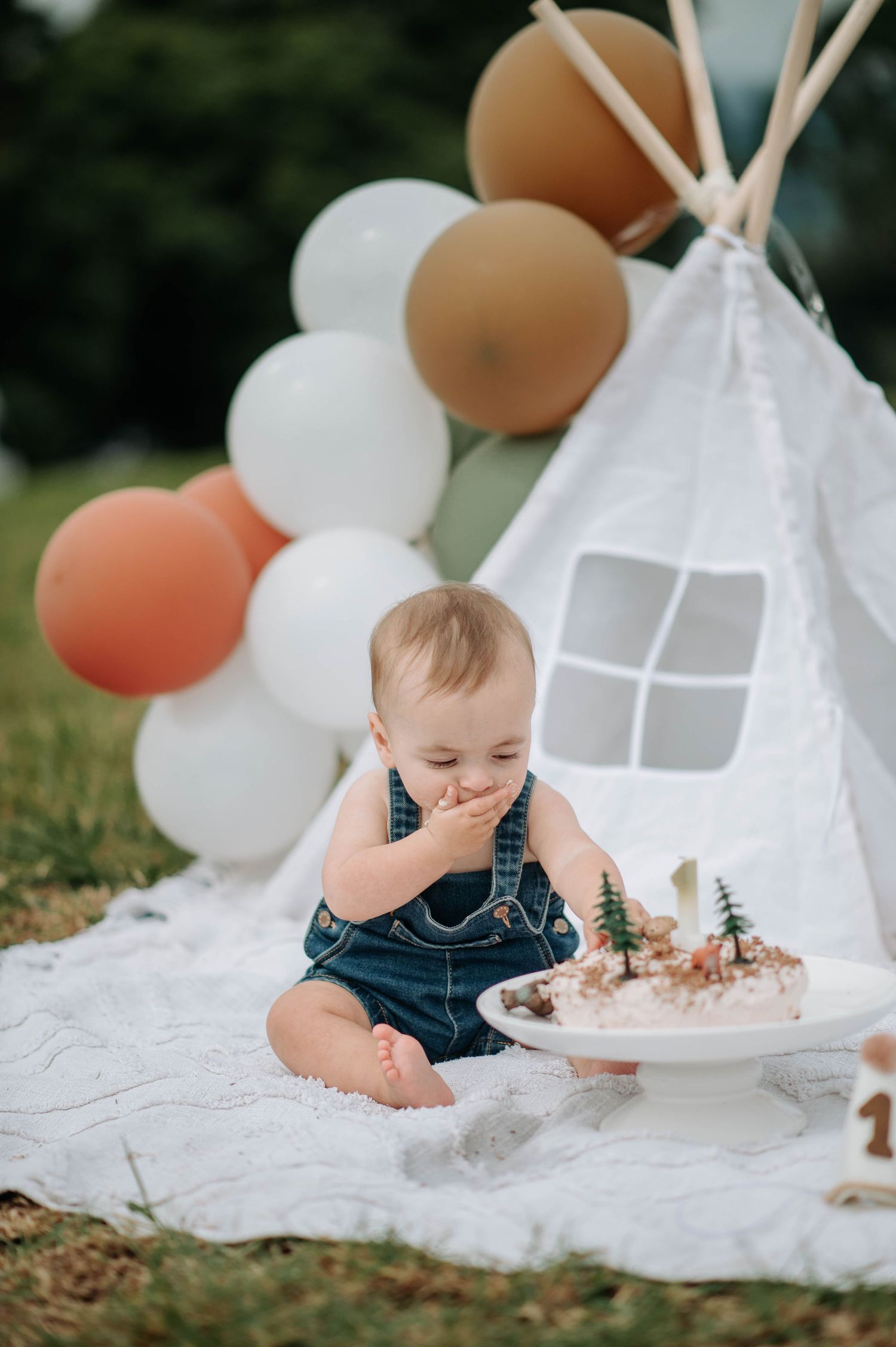 Baby in blue overalls tasting birthday cake beside white teepee and balloons.