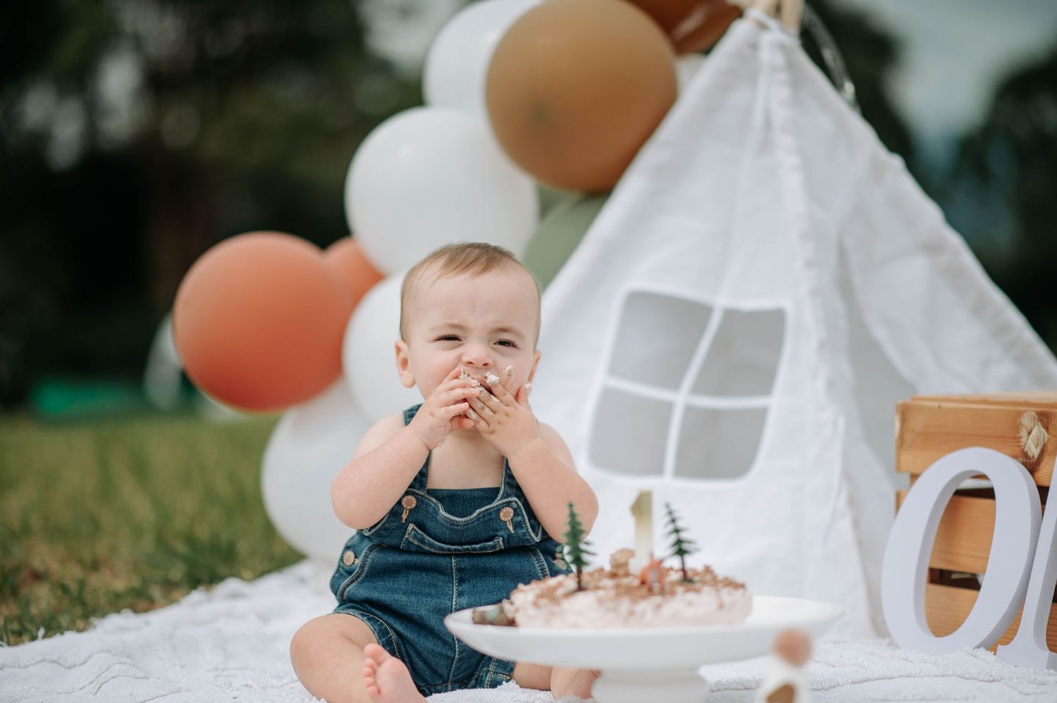 Baby giggling with hands over mouth next to birthday cake and white teepee tent.
