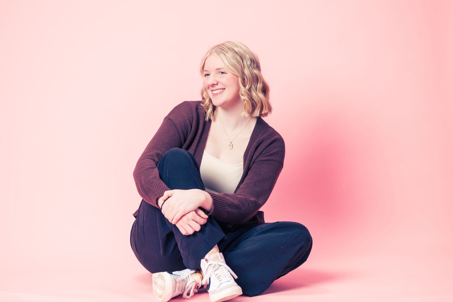 Smiling blonde woman sitting cross-legged on floor in purple cardigan and jeans against pink background.