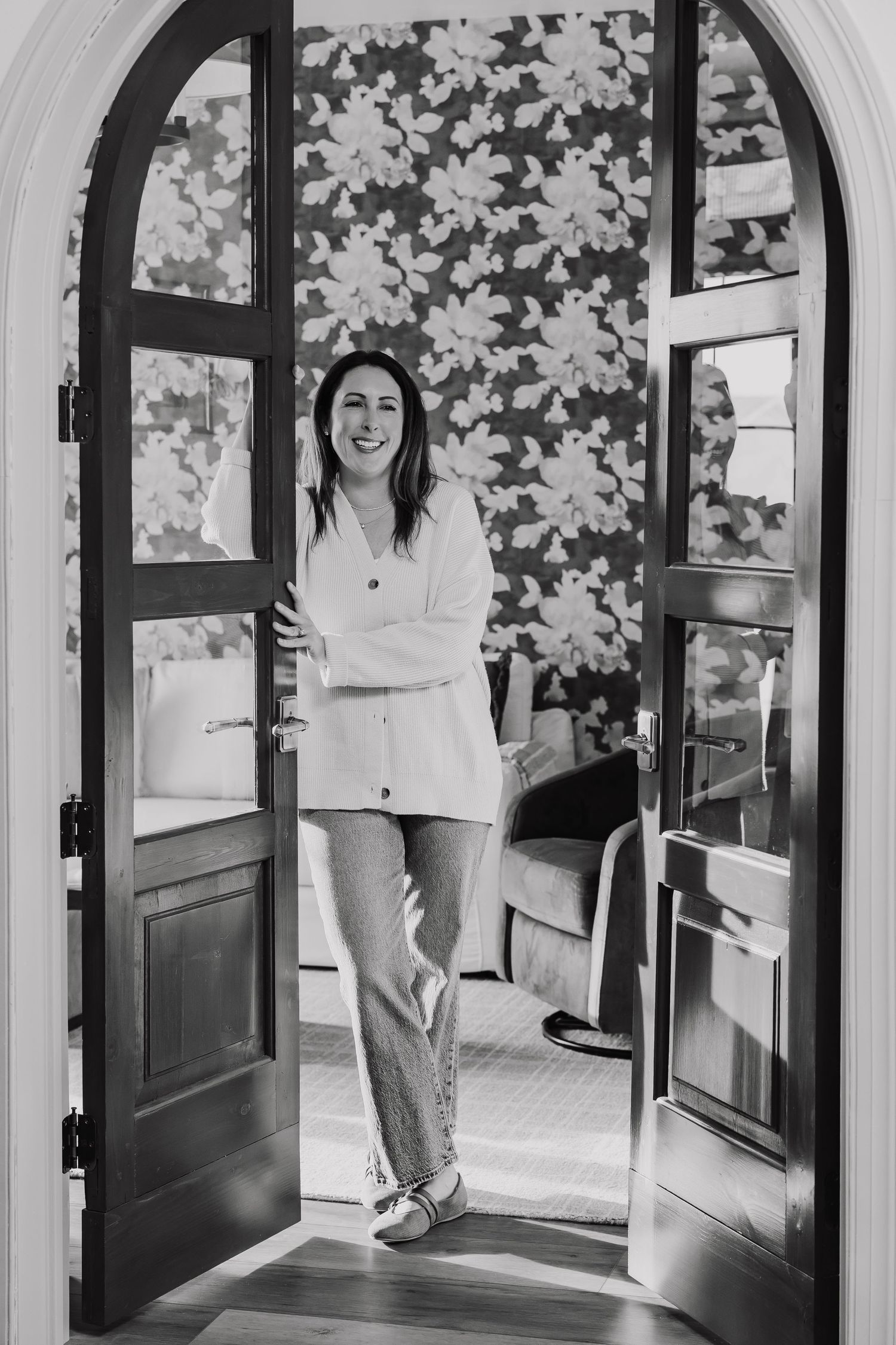Black and white photo of a smiling woman in jeans and white shirt standing in an arched doorway with floral wallpaper.