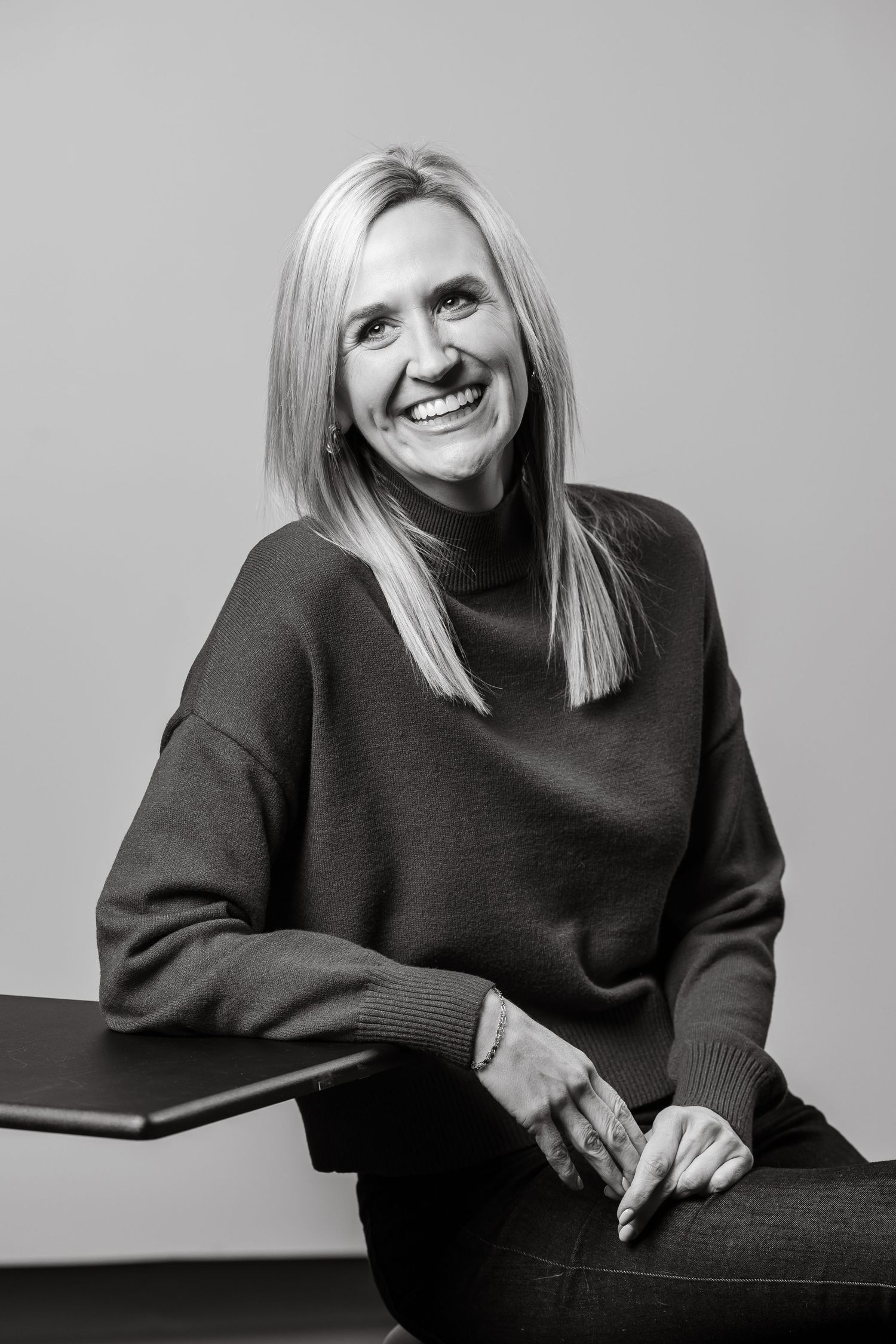 Smiling blonde woman in black sweater sits relaxed, black and white professional portrait photo.