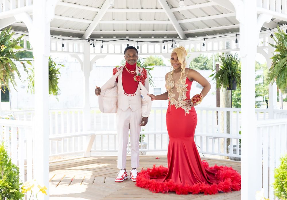 A couple poses for photos in a white gazebo, with the woman wearing a stunning red formal gown with feathers.