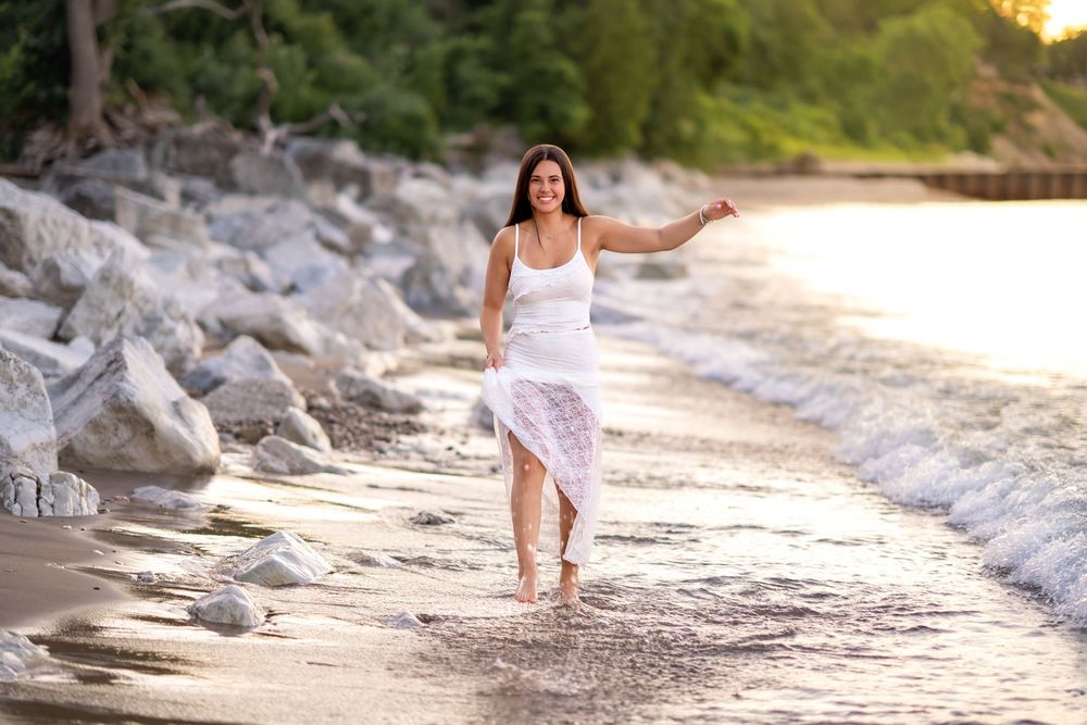 A person in a white dress walks along a rocky beach shoreline during golden hour, splashing in gentle waves.