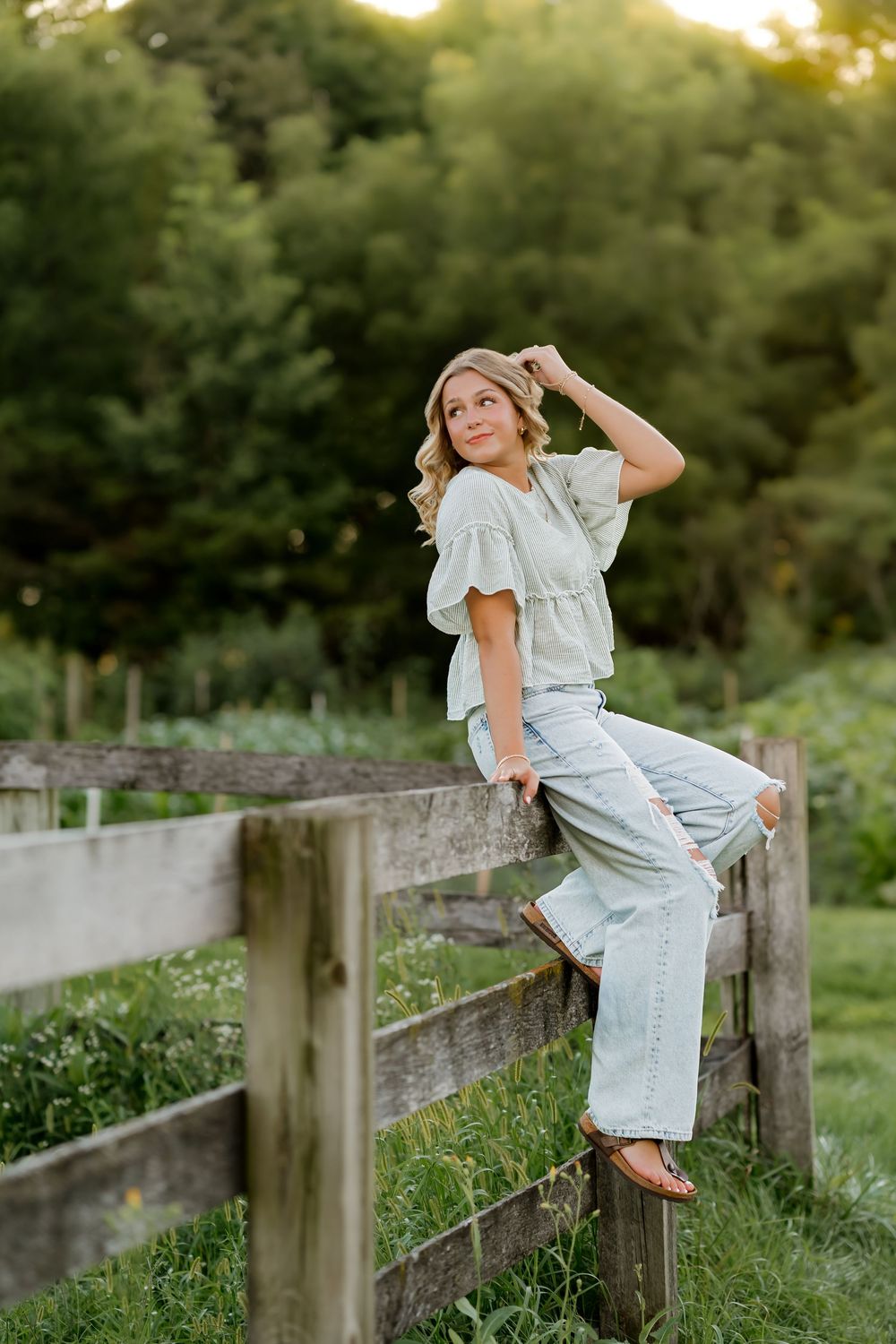 Young woman in light blue jeans and ruffled top sitting on a rustic wooden fence surrounded by lush greenery.