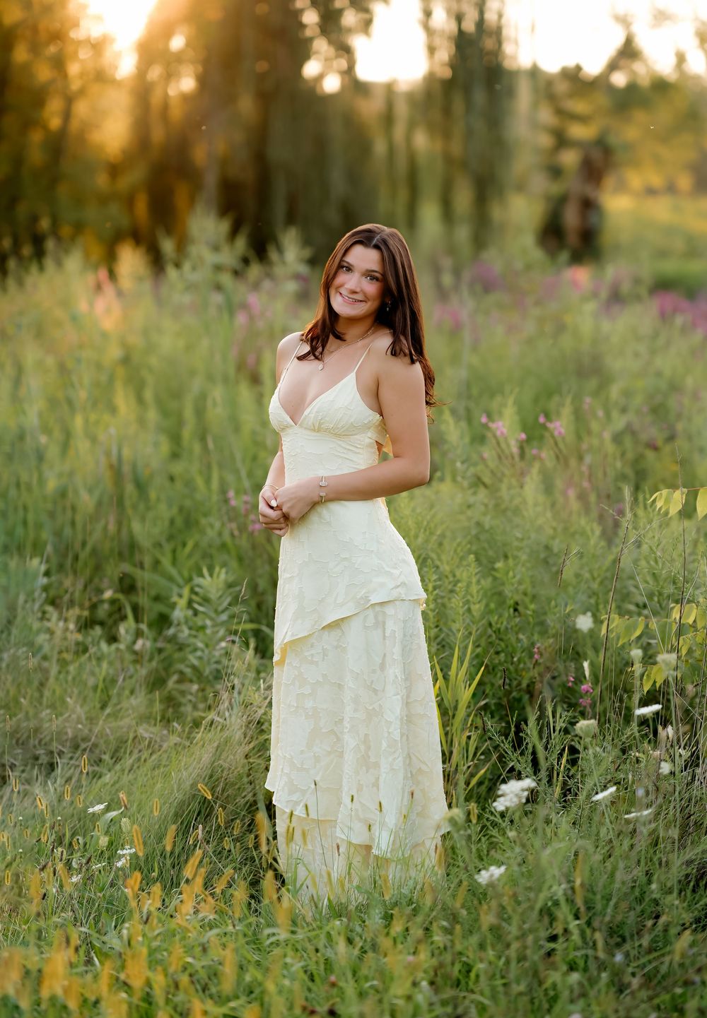 A young woman in a white lace gown stands gracefully in a wildflower meadow at golden hour.