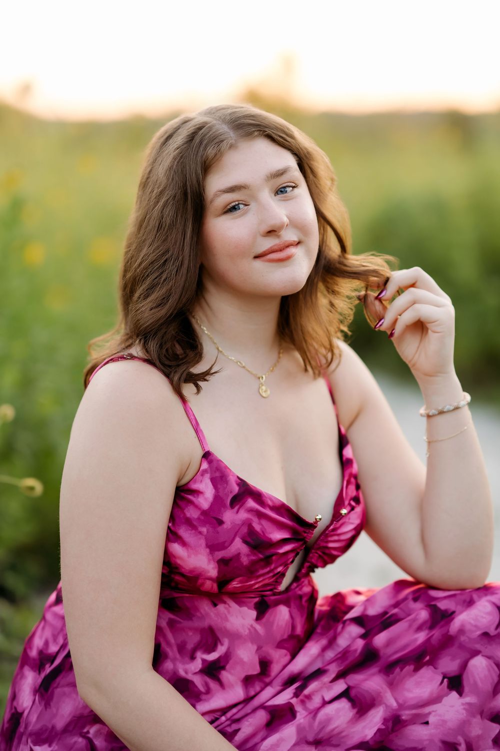 Young woman in pink floral dress sitting in a sunlit field, smiling softly while touching her hair.