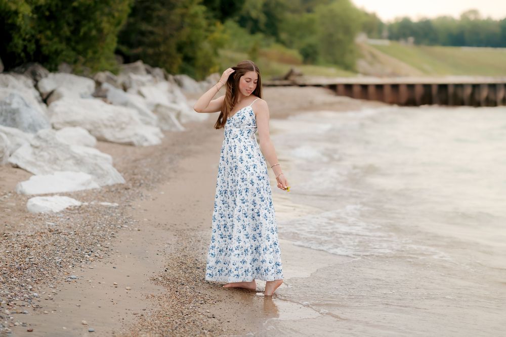 A young woman in a floral maxi dress walks barefoot along a sandy beach shoreline near rocky cliffs.