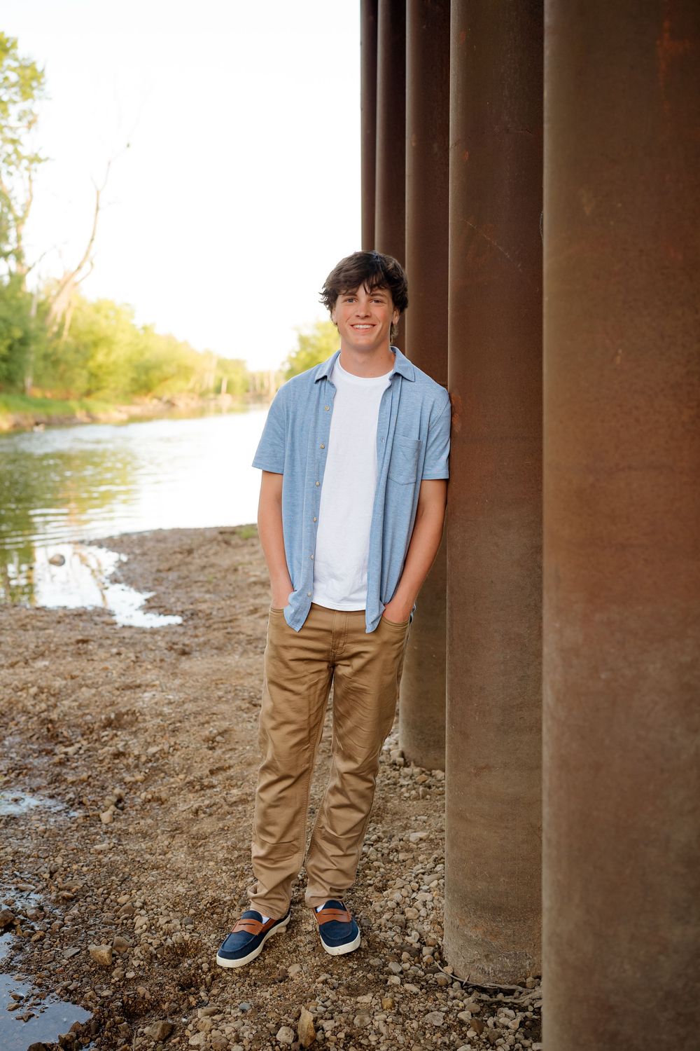 Smiling teenage boy in blue shirt and khakis leaning against rusty bridge pillar by a river.