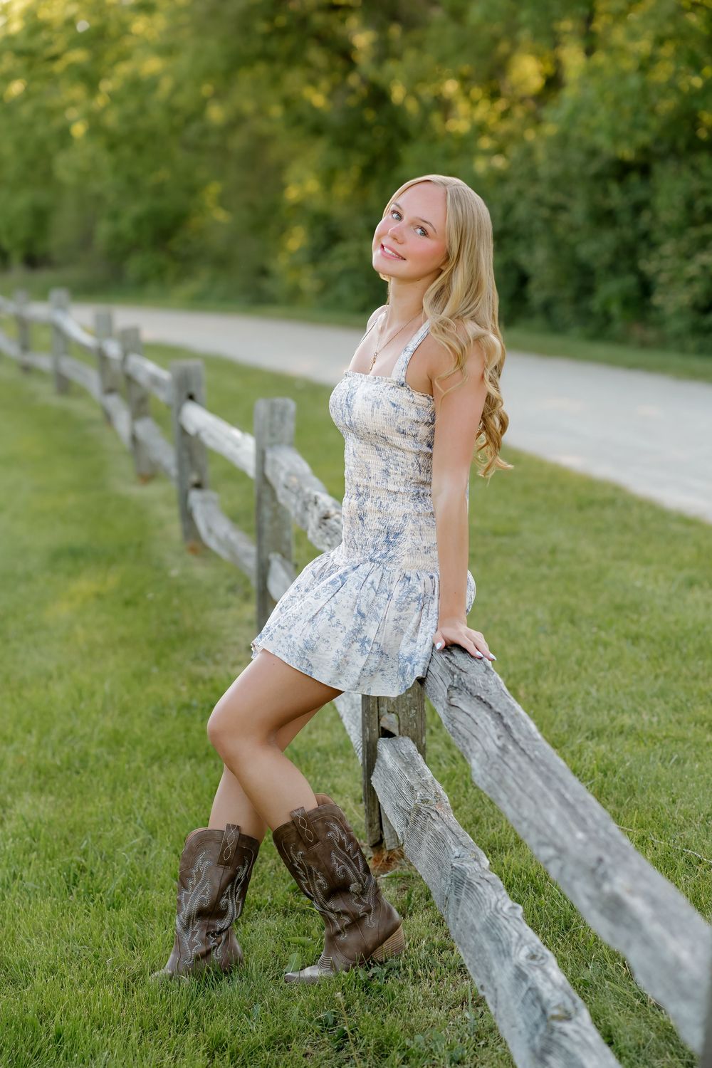 Young blonde woman in floral dress and cowboy boots leaning on rustic wooden fence along a tree-lined country road.