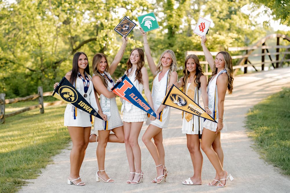 Five young women in white outfits holding college pennants on a tree-lined path celebrating their university acceptances.