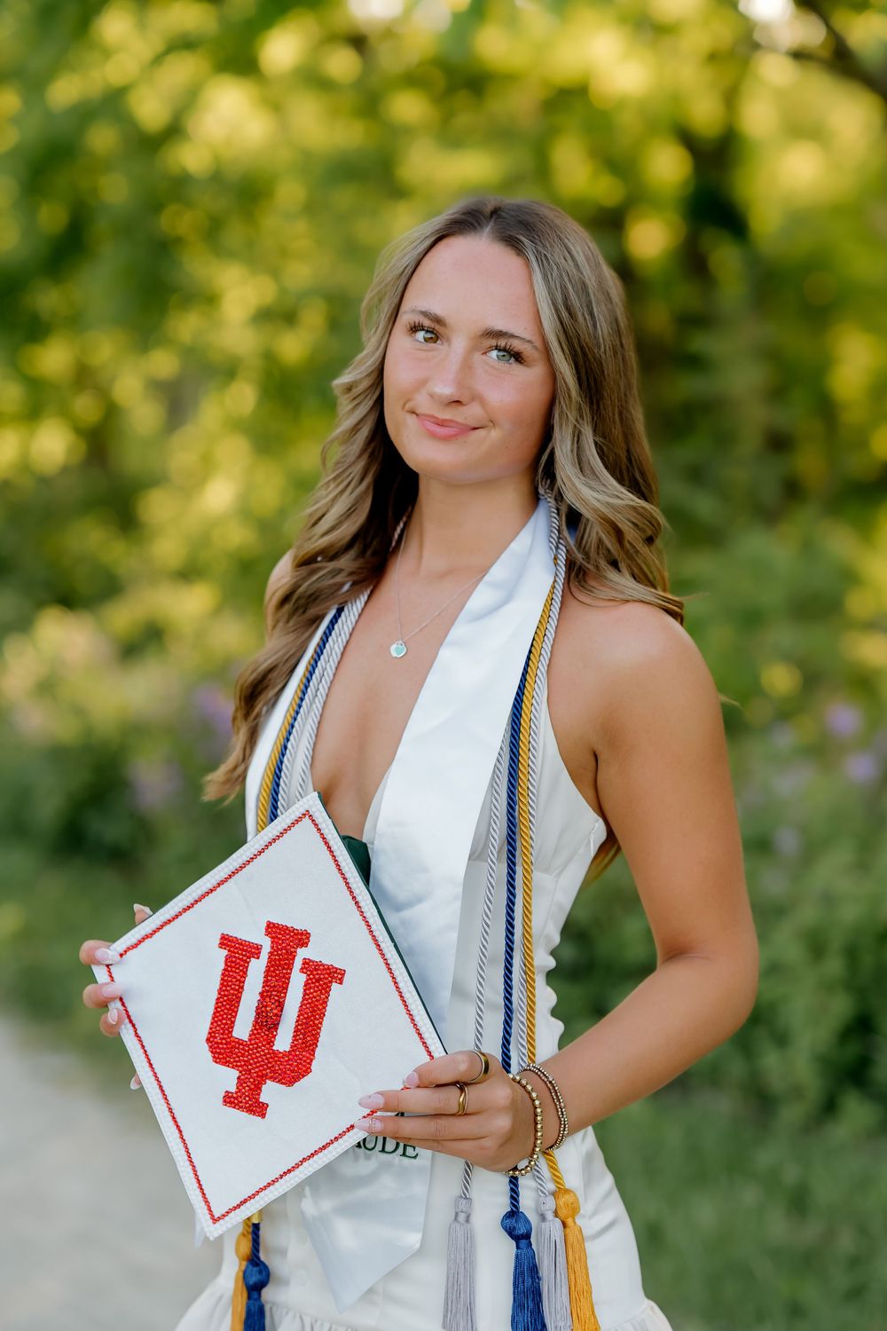 A smiling young woman in graduation attire holds an Indiana University cap outdoors among lush green trees.