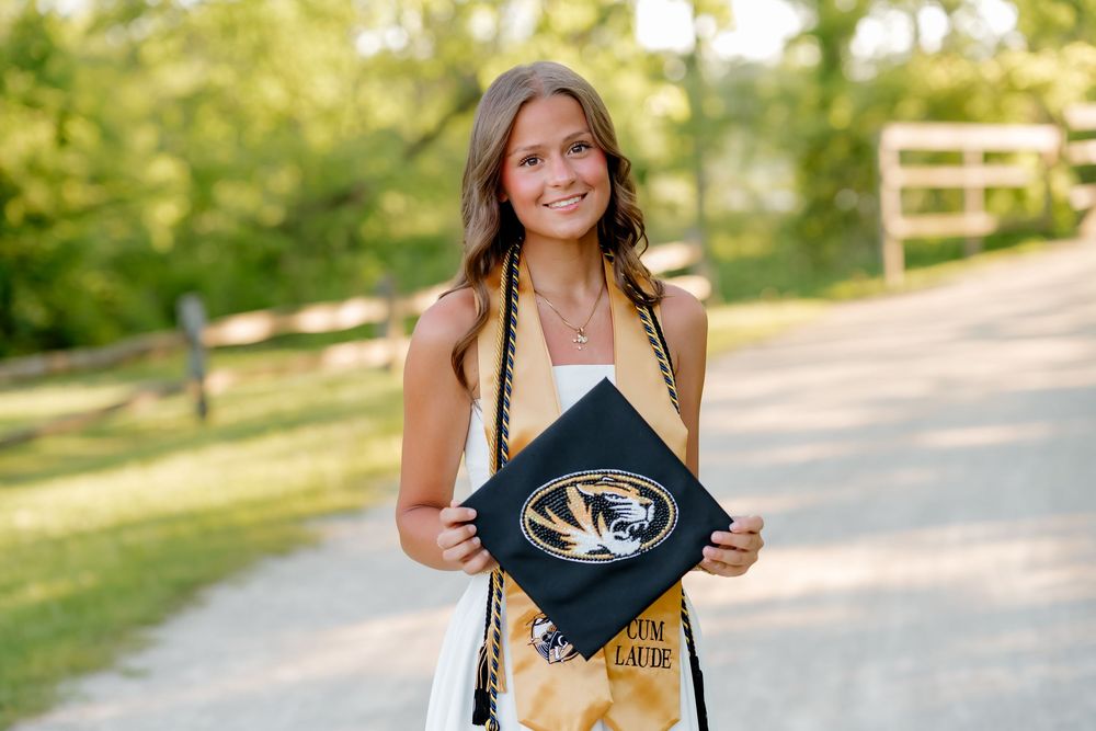 Smiling graduate holding a University of Missouri cap with gold and black tassels outdoors.