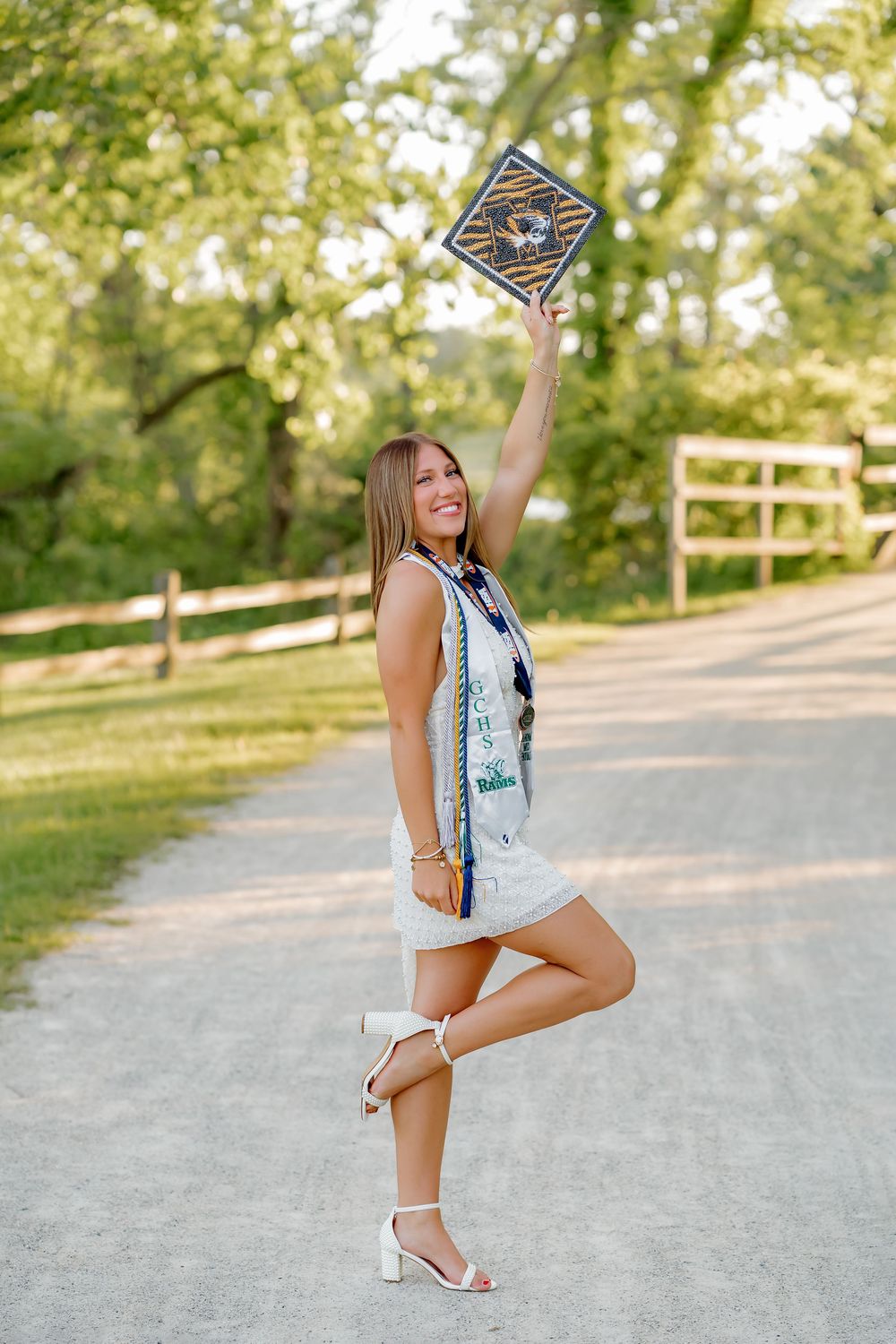 Smiling graduate in white dress holds cap overhead on a sunny tree-lined path, wearing honor cords and sash.