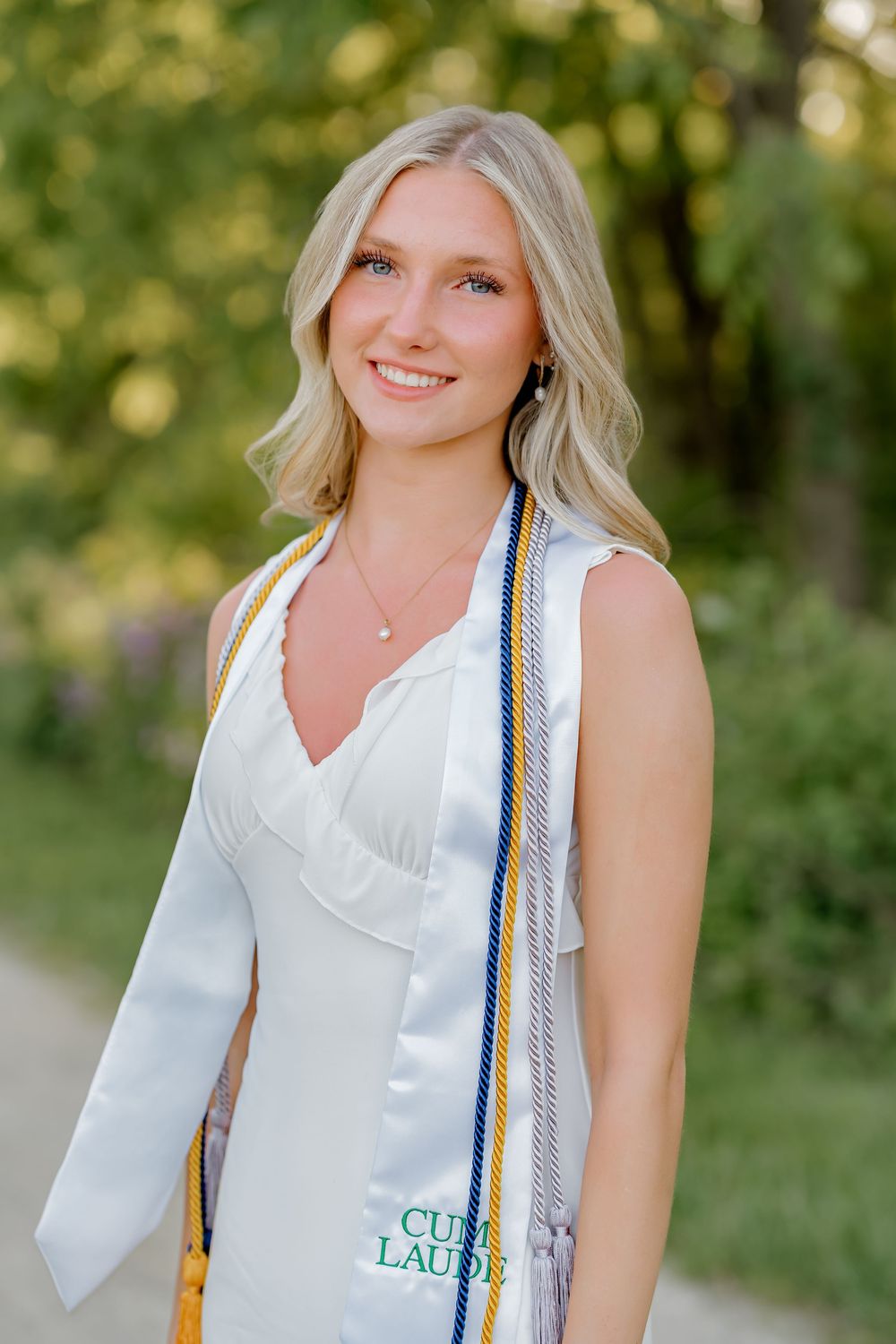 Blonde graduate smiles outdoors wearing white gown with Cum Laude stole and blue and gold honor cords.