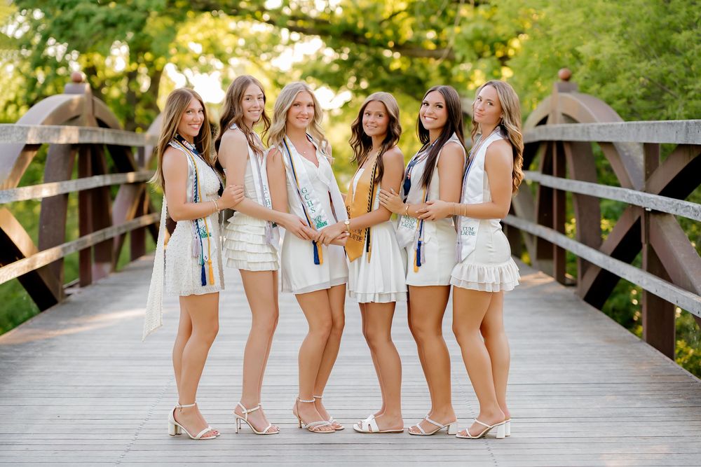 Six young women in white dresses pose together on a wooden bridge surrounded by lush green trees.
