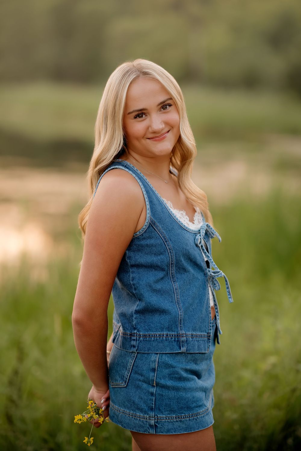 A smiling blonde teenage girl in a denim romper holds wildflowers in a sunlit outdoor field near water.