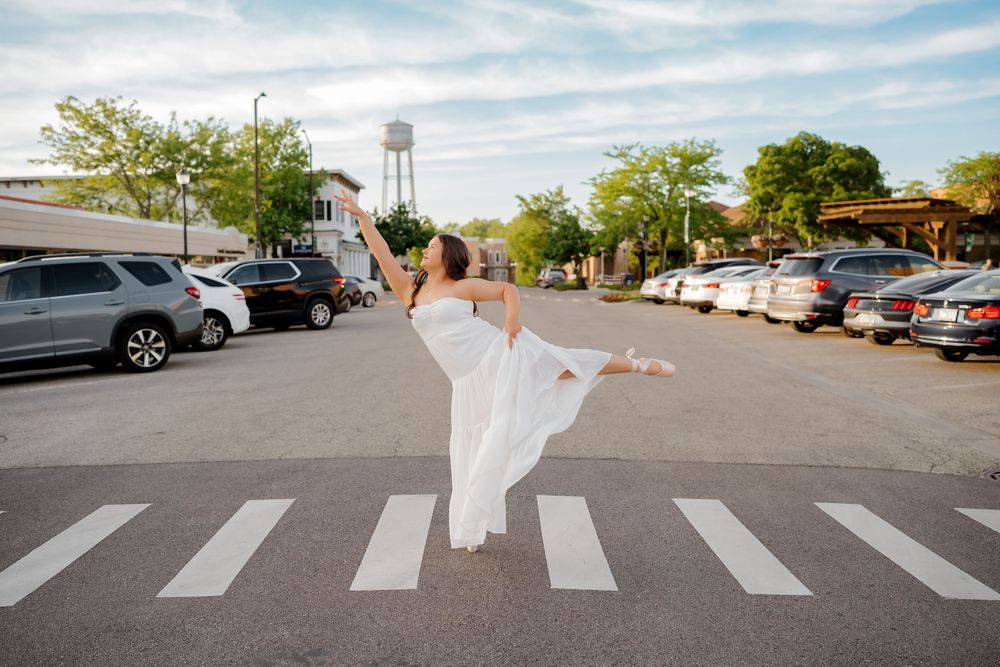 A dancer in a white dress performs an elegant arabesque pose at a crosswalk in an urban street setting.